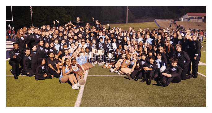 A large group of people in coordinated uniforms and costumes are gathered on a sports field around several trophies, posing for a celebratory group photo. Description generated by AI