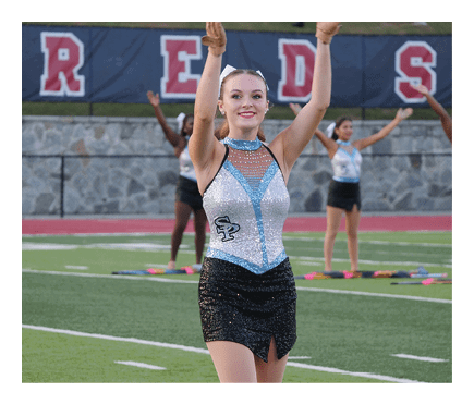A person in a sparkly blue and black performance outfit is standing on a sports field with arms raised while other people in similar attire perform in the background. Description generated by AI