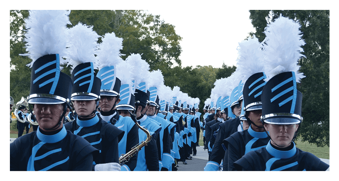 A marching band dressed in blue and black uniforms with tall hats featuring white plumes is lined up outdoors in formation. Description generated by AI