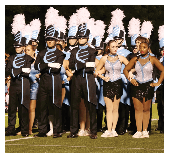 People in marching band uniforms and sequined dance costumes stand in formation on a field at night. Description generated by AI