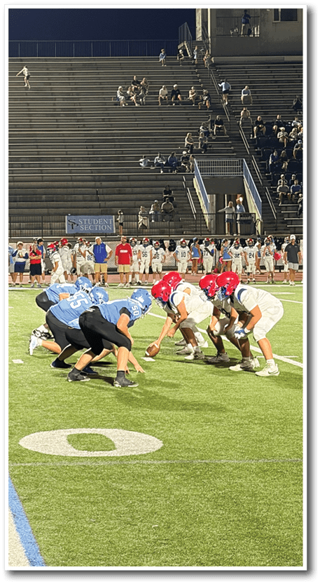 Players in blue and white uniforms are lined up on a football field facing each other as a game is about to start under stadium lights with empty bleachers in the background. Description generated by AI