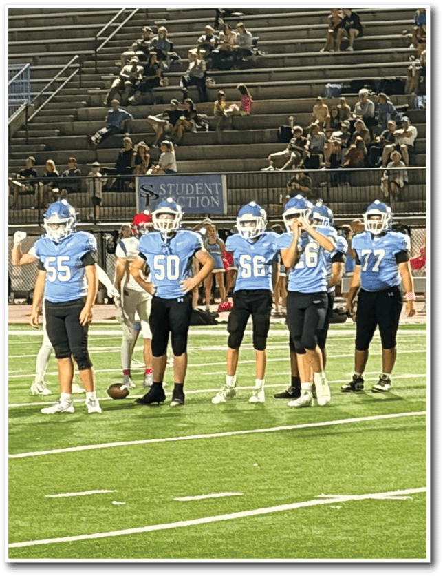 A group of people wearing blue and white football uniforms are standing on a green field with spectators seated in the bleachers behind them. Description generated by AI