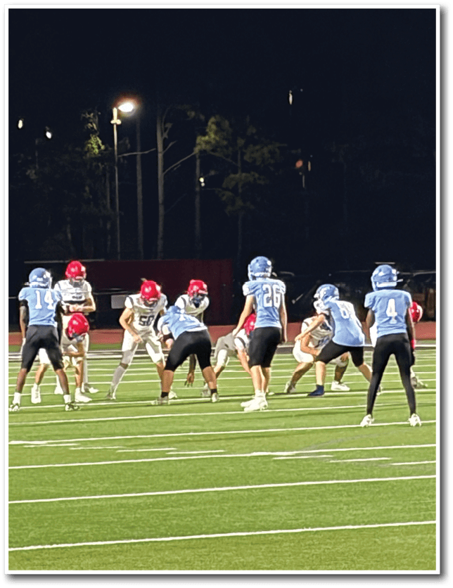People wearing helmets and sports uniforms are lined up on a football field at night preparing for a play. Description generated by AI