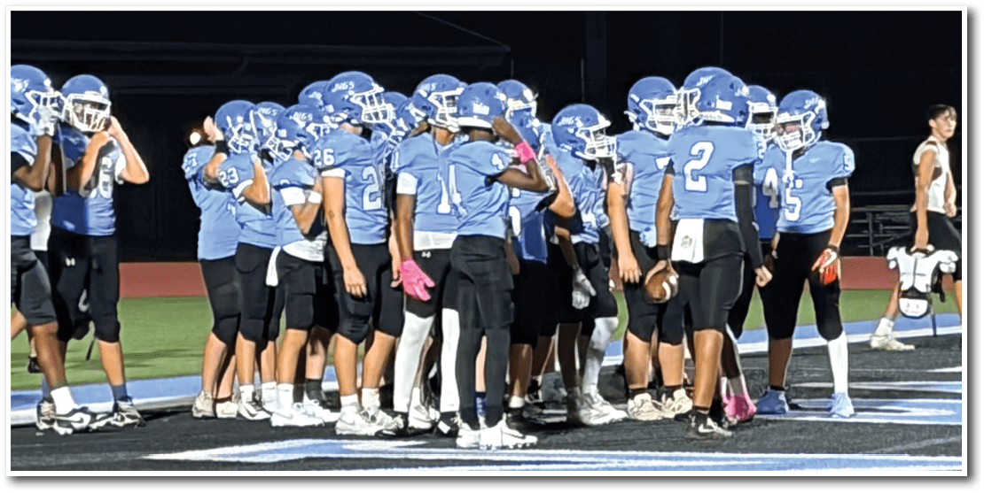 A group of people in blue sports uniforms and helmets are huddled together on a football field at sunset. Description generated by AI