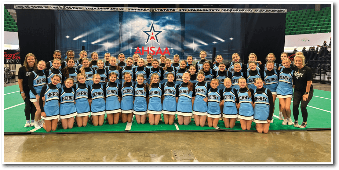 A large group of people dressed in matching blue and black cheerleading uniforms pose together on a green floor in front of a backdrop with the AHSAA logo. Description generated by AI