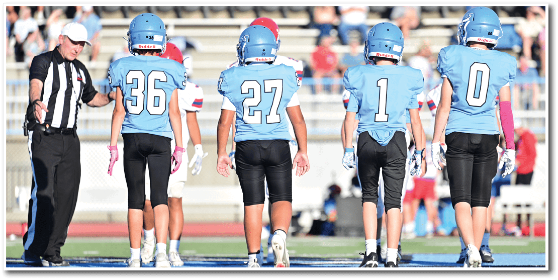 Four people in blue football uniforms and helmets stand on a field facing away from the camera with a referee gesturing beside them. Description generated by AI
