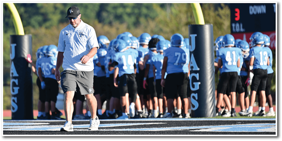 An adult wearing a white shirt, shorts, and a cap walks on a football field while a group of people in blue jerseys and helmets stand together near goalposts labeled “JAGS." Description generated by AI