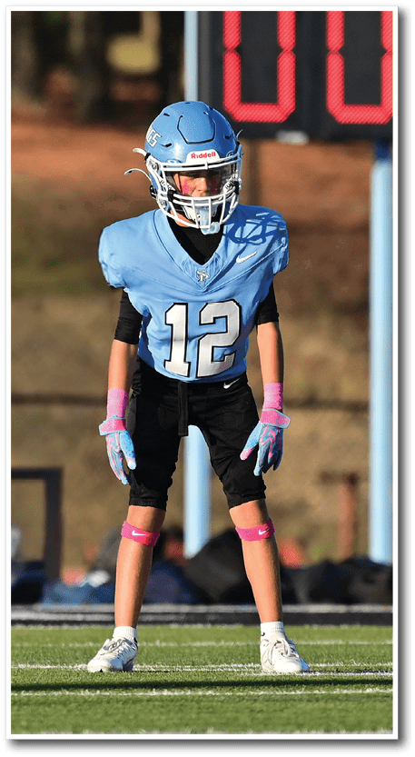 Two children wearing football uniforms and helmets stand on a field with a scoreboard in the background. Description generated by AI