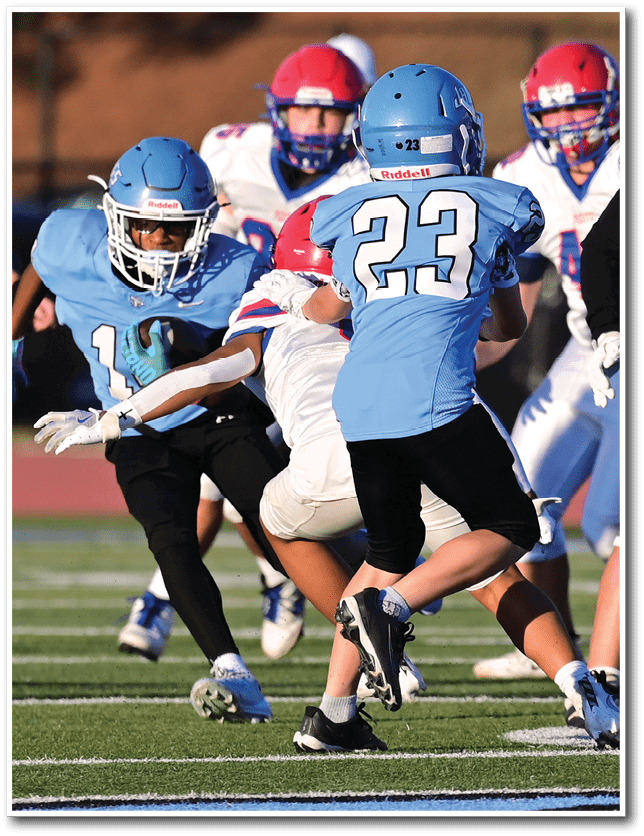 Several people wearing blue and red football uniforms are actively engaged in a play on a grassy field during a game. Description generated by AI