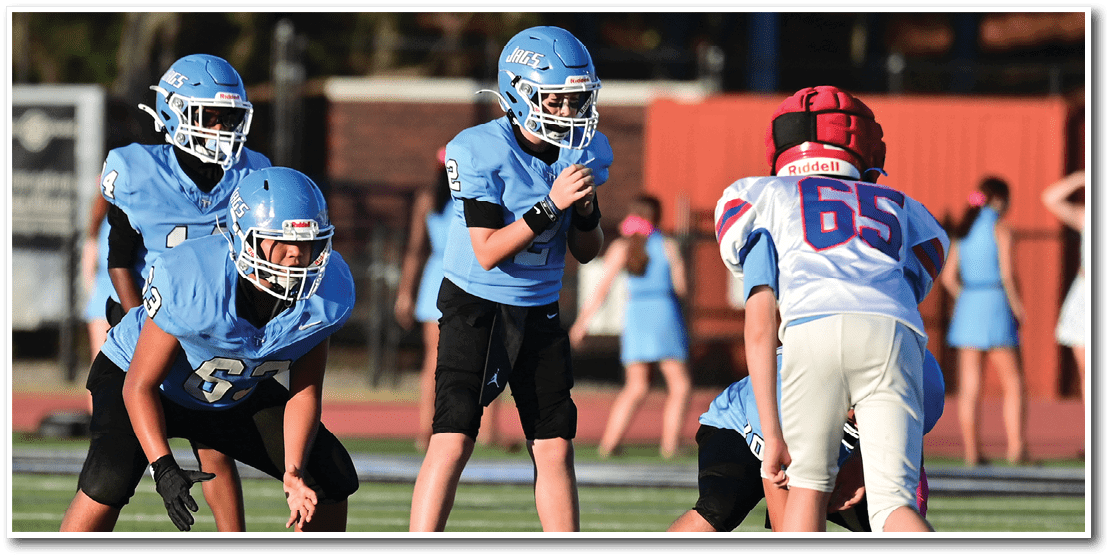 Children in football uniforms are positioned on a field preparing for a play during a game. Description generated by AI