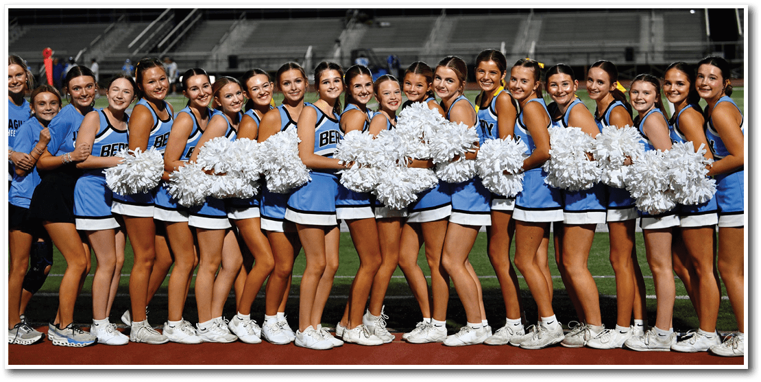 A group of adults in blue and white cheerleading uniforms stand closely together on a sports field at dusk, smiling and holding white pom-poms. Description generated by AI