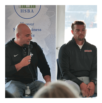 Two people wearing dark long-sleeve shirts are seated, with one holding a microphone and speaking while the other listens quietly. Description generated by AI