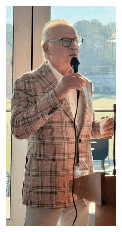 A senior wearing glasses and a plaid blazer is speaking into a microphone while standing behind a podium in a room with large windows showing an outdoor field. Description generated by AI