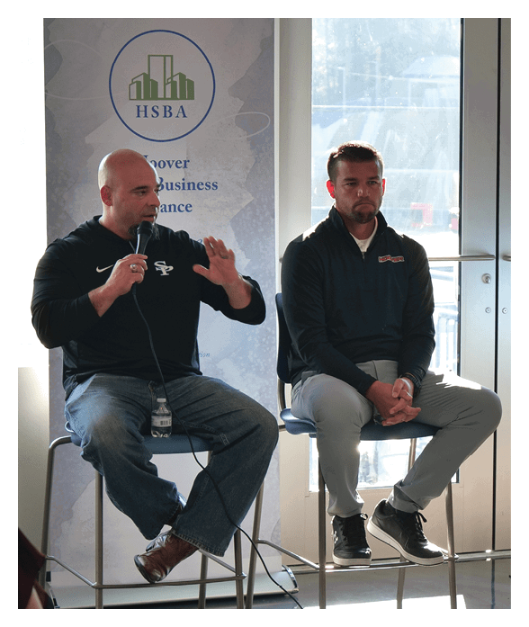 Two adults sit on high stools in a bright room with large windows, one speaking into a microphone while gesturing with a hand and the other sitting with hands clasped. Description generated by AI