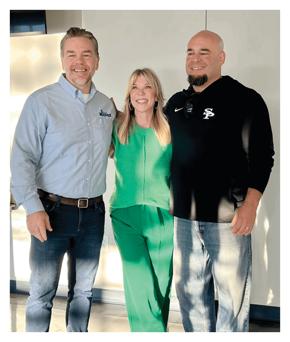 Three adults stand closely together smiling indoors with one wearing a light blue button-up shirt, another in a green sweater, and the third in a black hoodie with a logo. Description generated by AI
