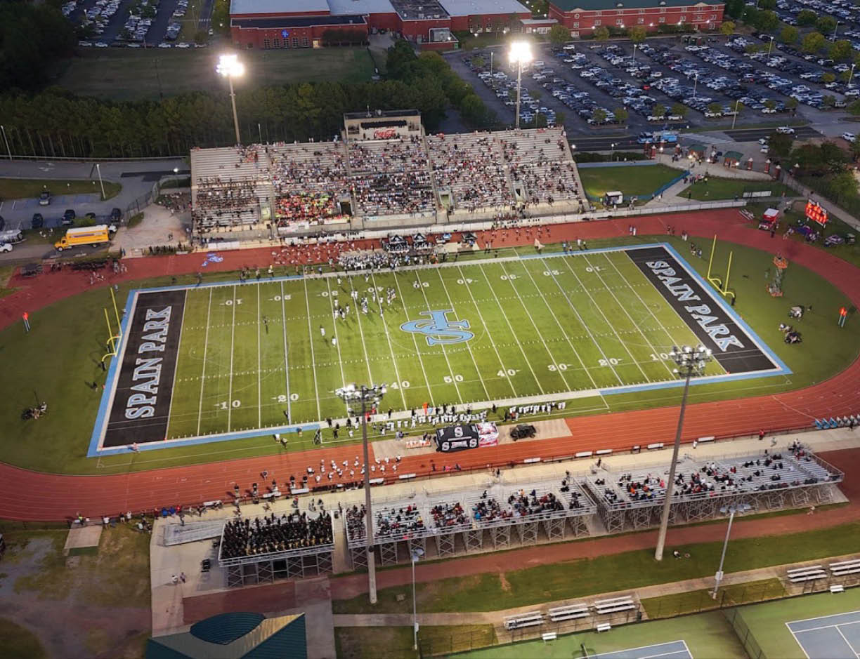 A football field with players on it is surrounded by a running track and bleachers filled with spectators under bright stadium lights during an evening game. Description generated by AI