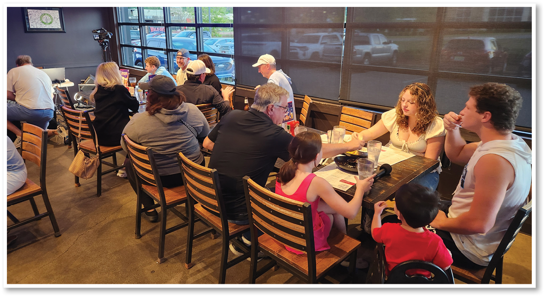 A group of people sitting around a long wooden table in a casual dining setting, engaging in conversation and sharing drinks. Description generated by AI