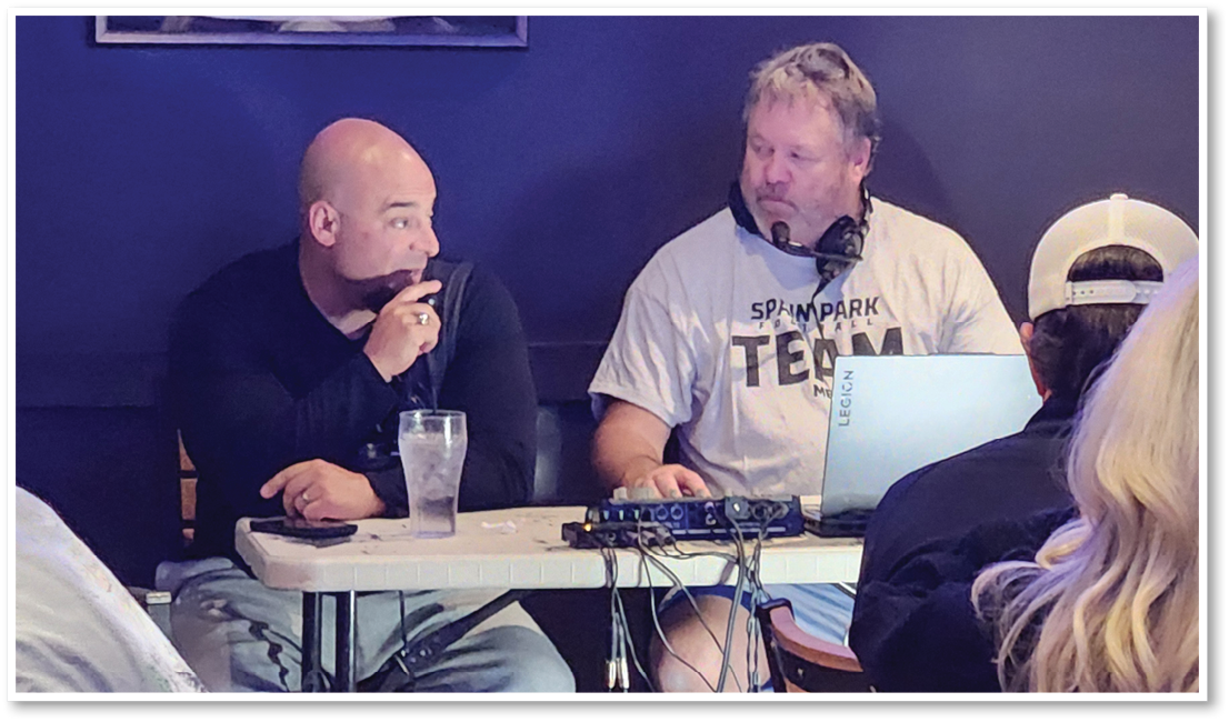 Two adults sit at a table with audio equipment and a laptop, one holding a microphone and the other wearing headphones, while several people sit nearby in a casual indoor setting. Description generated by AI