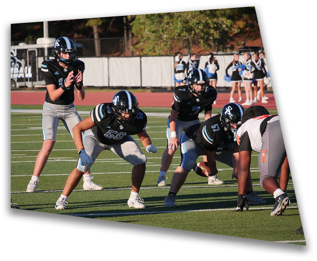A group of people wearing black and gray football uniforms are positioned on a football field, with one person standing behind the others in a quarterback stance. Description generated by AI