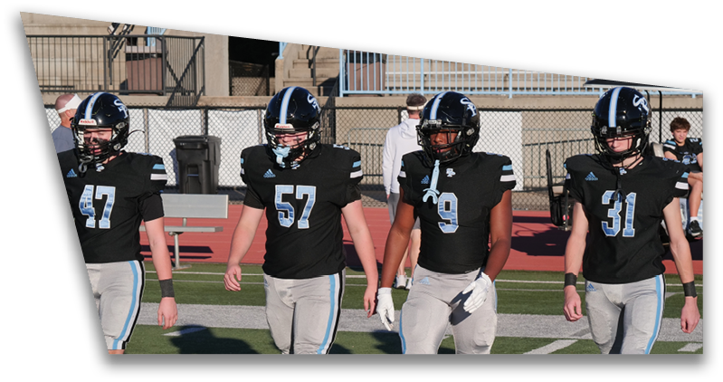 Four people wearing black football uniforms and helmets walk side by side on a football field with a fence and bleachers in the background. Description generated by AI