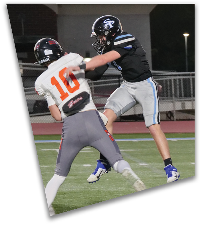 Two people wearing football helmets and uniforms are actively engaged in a play on a football field at night. Description generated by AI