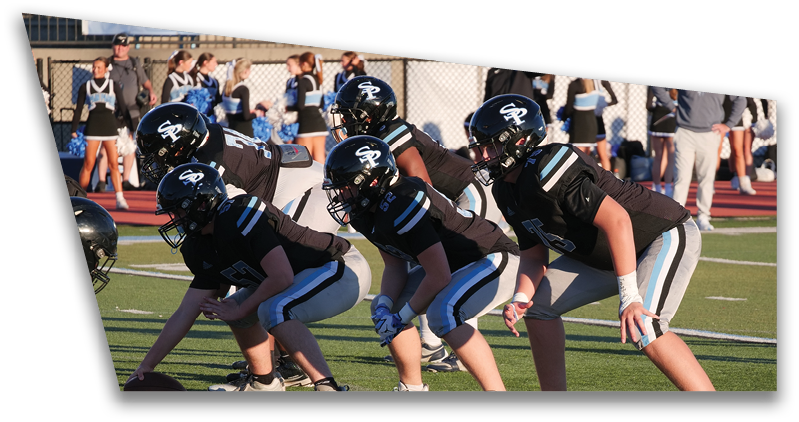A group of people in football uniforms are lined up on a field in a ready stance facing an opposing player holding a football. Description generated by AI
