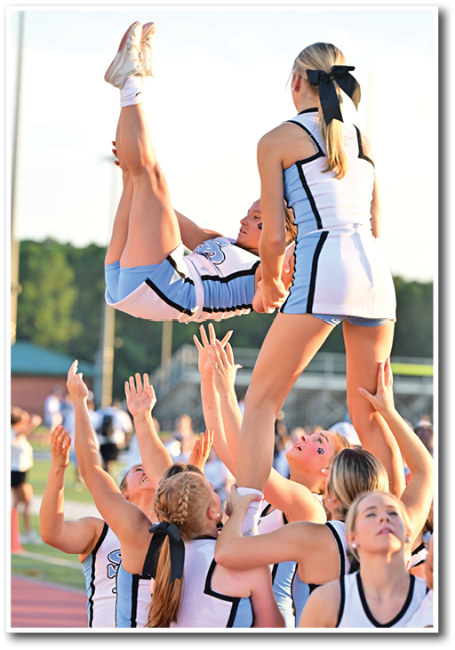 A group of people in cheerleading uniforms are performing a stunt outdoors on a sports field with one person being lifted and flipped in the air while others support and catch them. Description generated by AI