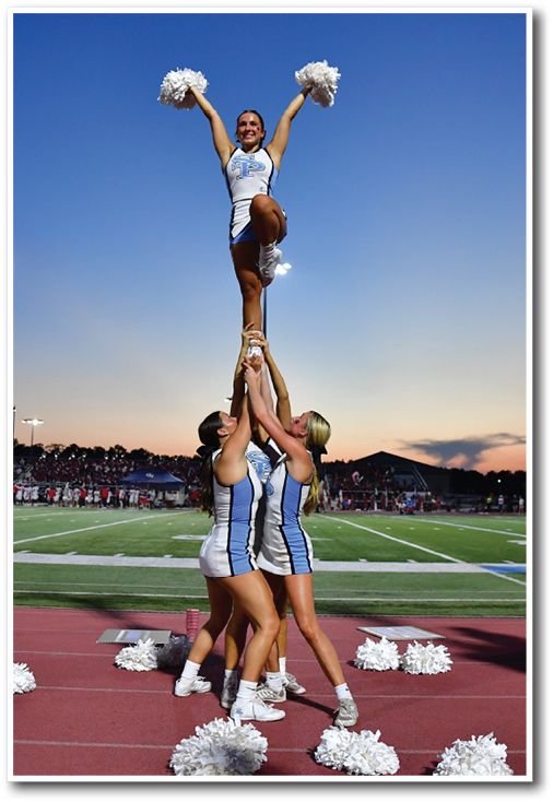 People in cheerleading uniforms perform stunts and hold pom-poms on the sidelines of a sports field during sunset. Description generated by AI