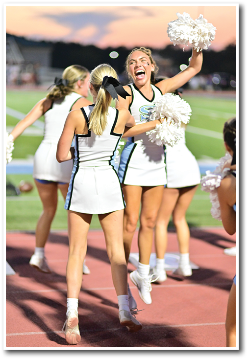 Three people in cheerleading uniforms are interacting outdoors with one person laughing joyfully and another with a black bow in their hair. Description generated by AI