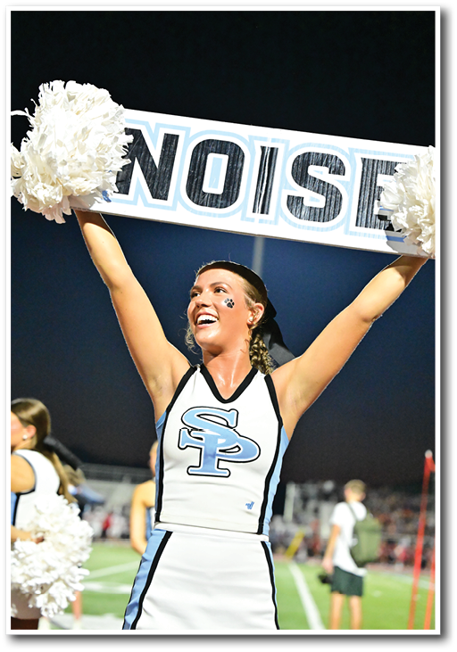 A person in a cheerleading uniform with “SP" on the front is smiling and holding up a sign that says "NOISE" with pom-poms on each side at a nighttime outdoor sports event. Description generated by AI