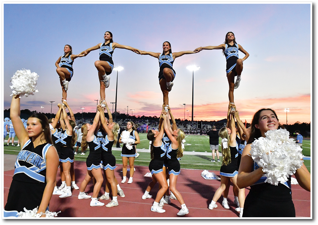 A group of cheerleaders in matching uniforms perform a coordinated stunt on a sports field at sunset with some holding pom-poms and others lifted in the air. Description generated by AI