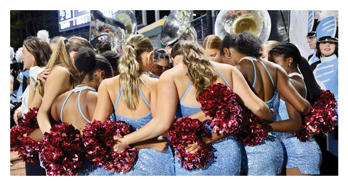A group of people dressed in sparkly blue outfits with red pom-poms stand closely together with their arms around each other in a huddle. Description generated by AI