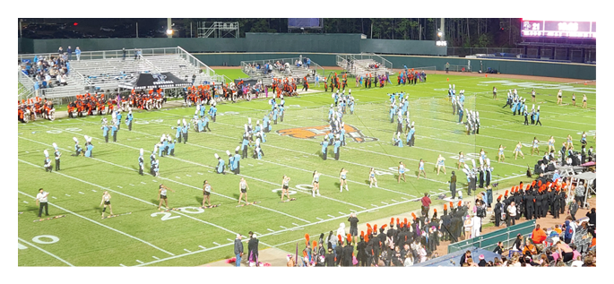 A marching band dressed in light blue uniforms is performing on a football field at night with a crowd watching from the stands. Description generated by AI