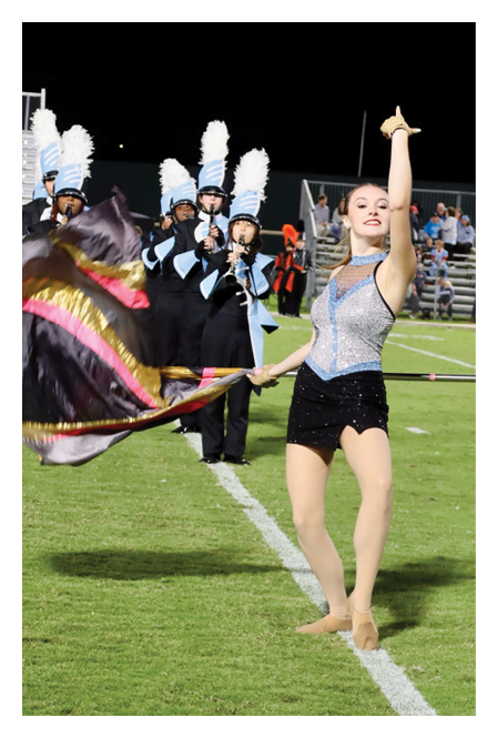 A group of people in marching band uniforms with tall white plumes are playing instruments on a field while a person in a sparkly outfit waves a colorful flag near the sideline. Description generated by AI