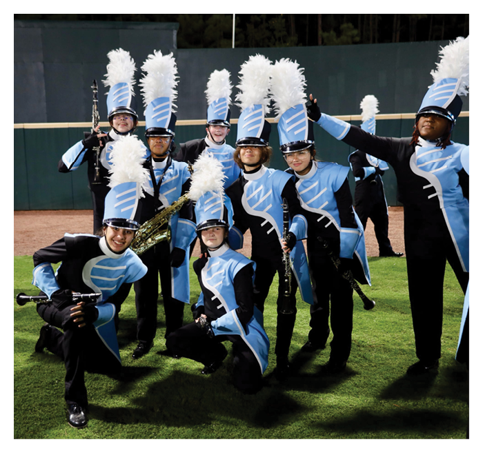People in blue and black marching band uniforms with tall hats and white plumes are posing on a grassy field, some standing and some kneeling, holding musical instruments. Description generated by AI