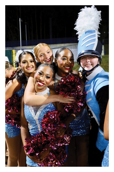 A group of smiling people dressed in sparkly blue dance costumes and one person in a blue marching band uniform with a tall white plume pose closely together holding red pom-poms. Description generated by AI
