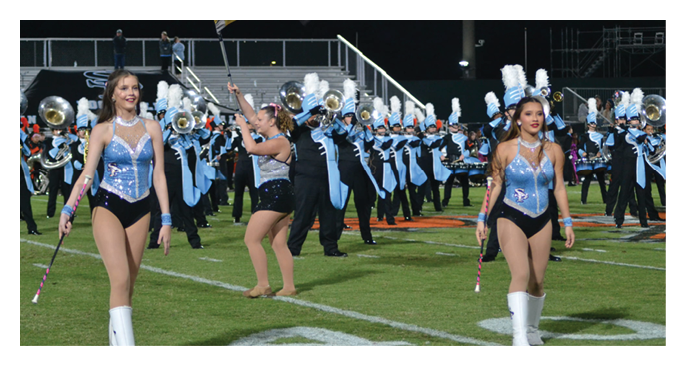 Two adults in blue and black performance outfits with white boots are twirling batons on a football field with a marching band playing instruments in the background. Description generated by AI