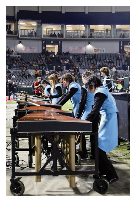 People wearing light blue uniforms are playing marimbas in a stadium with an audience in the background. Description generated by AI