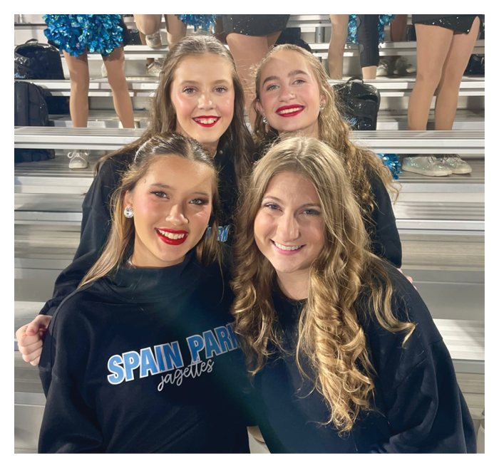 Four smiling people with long hair wearing black Spain Park Jolettes shirts pose closely together on metal bleachers with others standing in the background. Description generated by AI