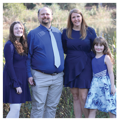 Four people dressed in blue clothing stand closely together outdoors with greenery in the background, smiling and posing for the photo. Description generated by AI