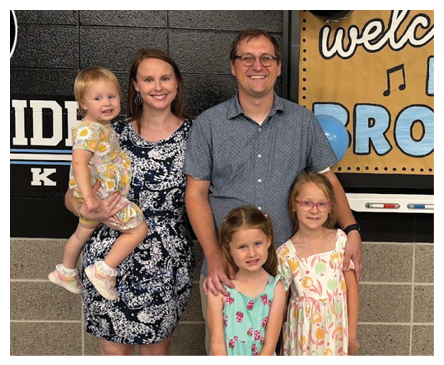 A group of five people, including three children in colorful dresses and two adults, stand together smiling in front of a wall with a sign and markers. Description generated by AI