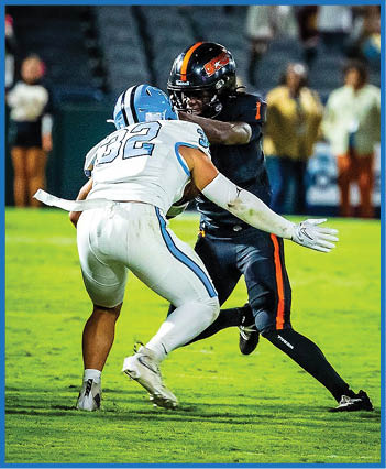 Two people in football uniforms are engaged in a play on a grassy field with blurred spectators in the background. Description generated by AI