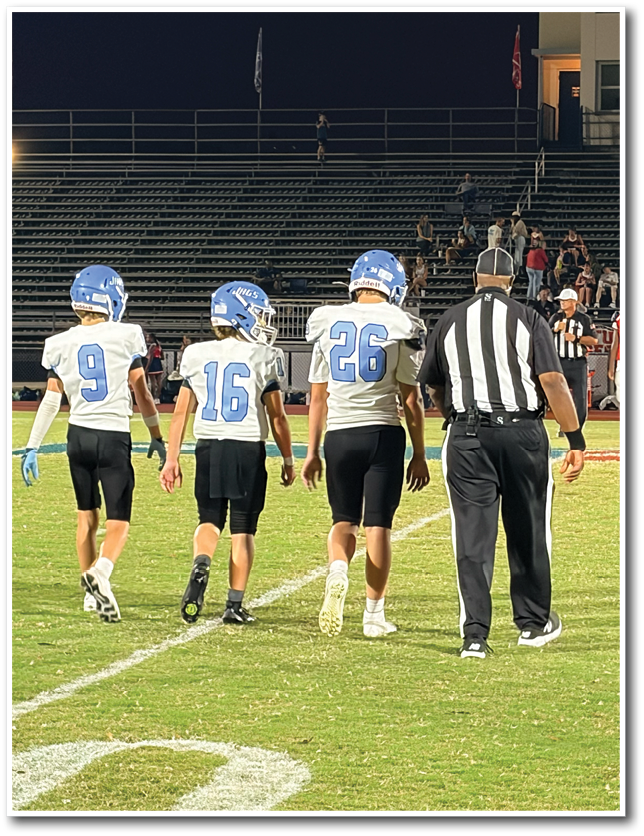 Four people, three in football uniforms and one in a referee uniform, walk side by side on a football field with empty bleachers in the background. Description generated by AI