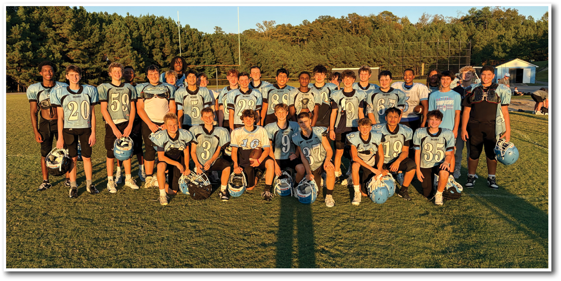A group of people wearing matching sports uniforms pose together on a grassy field with helmets in hand during daylight. Description generated by AI