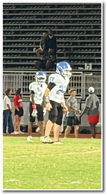 People in football uniforms stand on a field while others watch from bleachers under stadium lights at night. Description generated by AI