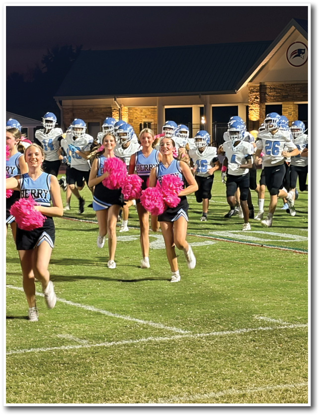 Three adults in cheerleading uniforms holding pink pom-poms run on a football field with a group of football players in helmets and uniforms running behind them at night. Description generated by AI