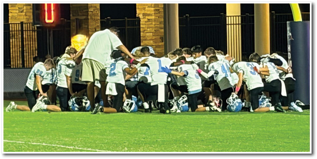 A group of people wearing sports uniforms kneel together on a grassy field in a huddle. Description generated by AI