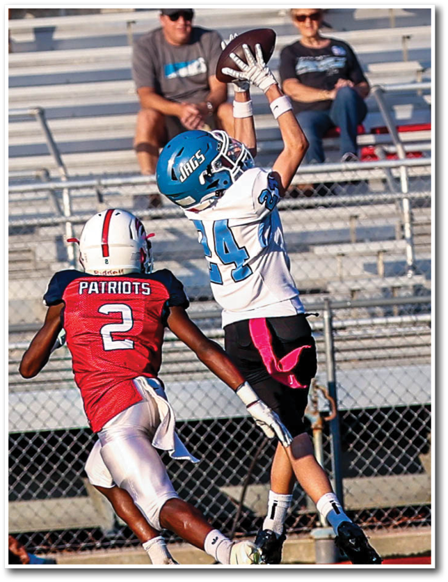 Two football players in action on a field with one player in a white and blue uniform jumping to catch the ball while the other player in a red and white uniform attempts to defend. Description generated by AI