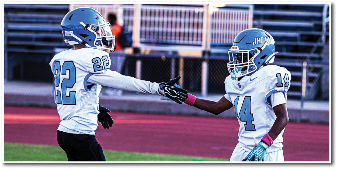 Two people wearing football uniforms are on a field, with one holding a football and the other reaching out to touch hands. Description generated by AI