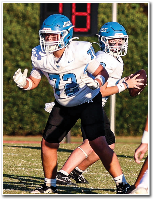 Two people in football uniforms are engaged in a play on a grassy field with one person holding a football and the other preparing to block an opponent wearing a red jersey. Description generated by AI